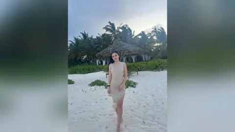 Elegant woman in a beige slip dress standing on a tropical beach with palm trees at dusk.