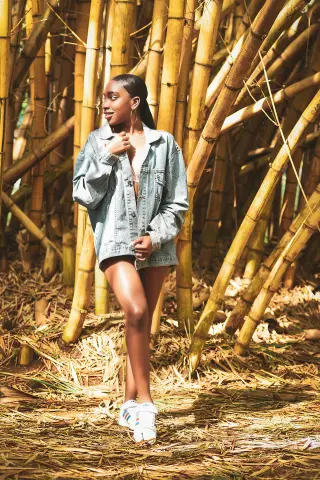 Stylish woman in denim jacket and shorts posing in a sunny bamboo forest setting.