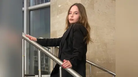 Young woman in a professional black blazer posing by a modern glass staircase in a bright office setting.