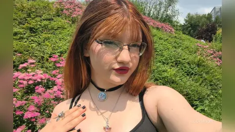LucyMoon is a redhead with glasses wearing a black tank top and a choker, posing outdoors in front of pink flowers.