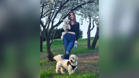 Smiling woman in black crop top and jeans holding a small white dog in a lush green park setting.