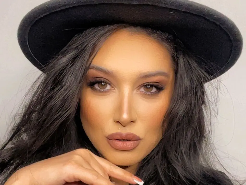 AnnaBanks poses wearing a black hat with her long dark hair framing her face and heavy eye makeup.