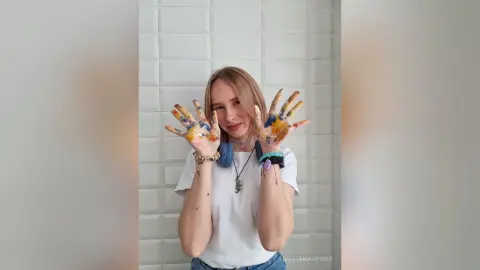 LisetteMaupredi poses with her hands up, showing paint splatters on her palms and fingers, wearing a white t-shirt and blue headphones.
