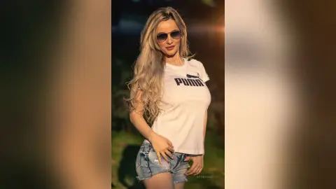 Blonde woman in a white Puma t-shirt and denim shorts posing outdoors in warm sunset lighting.