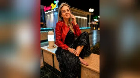 DelliaSmith sits on a stone ledge wearing a red jacket and a black ruffled skirt, posing with one bare foot visible.