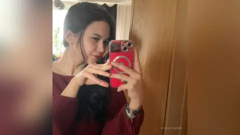Young woman in a cozy burgundy sweater taking a mirror selfie in a warm indoor setting.