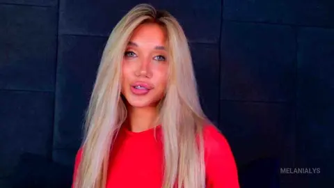 Blonde woman in a red long-sleeve top posing with a soft expression against a dark textured background.