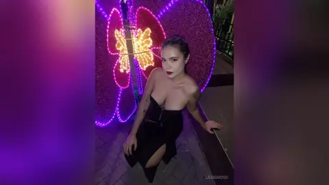 Glamorous woman in a black cutout dress posing before vibrant purple neon butterfly lights at night.