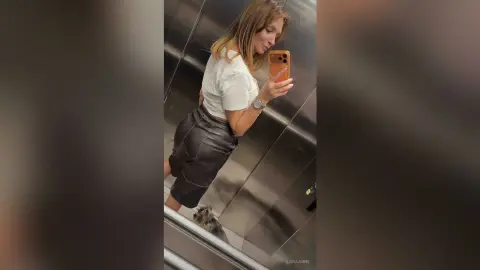 Woman in white t-shirt and black leather trousers taking a mirror selfie in a modern elevator.