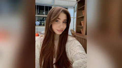 Smiling young woman with long brown hair wearing a white lace blouse in a bright, cozy indoor setting.