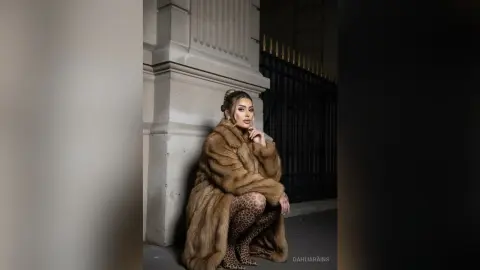 Elegant woman in a luxurious brown faux fur coat and leopard print leggings posing by a classic stone building.