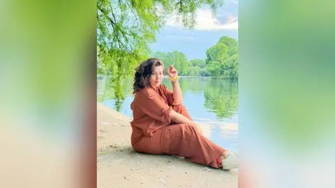CleoLangley sits on a concrete ledge by a calm lake, wearing a brown short-sleeved top and matching long skirt with her hair down.