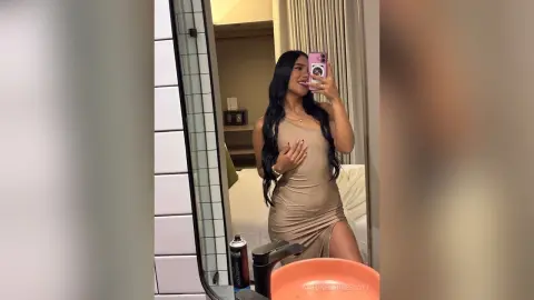 Smiling woman in a beige bodycon dress taking a mirror selfie in a modern bathroom.