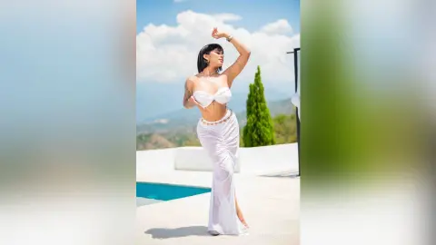 Stylish woman in a white bikini and maxi skirt posing by a luxury pool under a sunny summer sky.