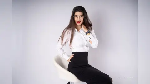 Smiling brunette woman in a white button-down shirt and black skirt sitting in a bright studio setting.