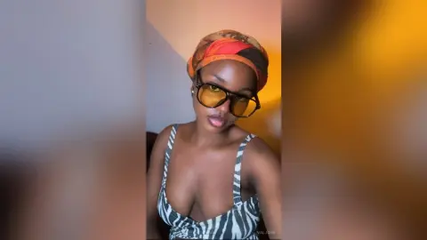 Valjohn wears a patterned headwrap and large sunglasses while posing in a striped bikini top.