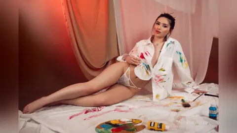Young woman in white shirt and lace lingerie reclining on a bed with paint palettes in a warm studio setting.