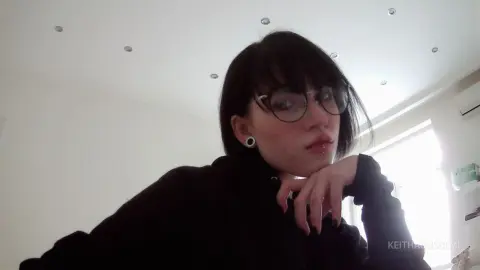 Moody portrait of a young woman with dark bangs and glasses wearing a black sweater in a bright room.