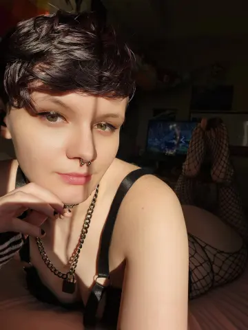 BarelyOlivia wears a black strap and silver chain necklace, posing with short dark hair and a nose ring in a dimly lit room.