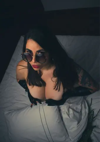 Moody portrait of a brunette woman in black gloves and sunglasses lounging on a white bed.