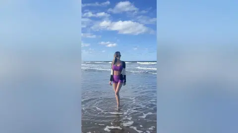 Young woman in purple swimwear standing on a sunny beach under a blue cloudy sky.