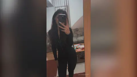 Young woman in a chic black long-sleeve outfit taking a moody mirror selfie in a modern room.