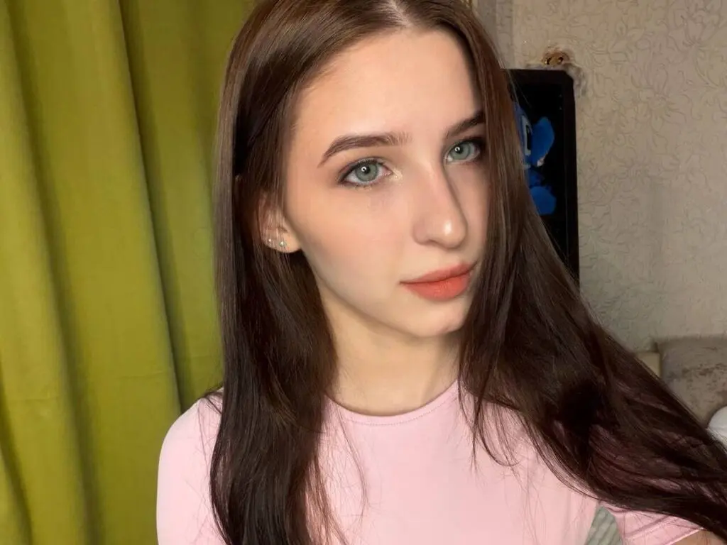 WanitaCruson has long brown hair and light eyes, wearing a pink t-shirt while posing in front of a green curtain.