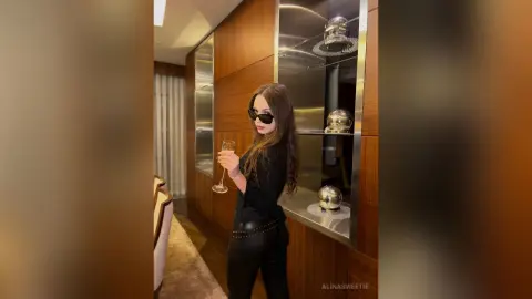 Stylish woman in black outfit and sunglasses holding a champagne glass in a modern luxury hotel room.