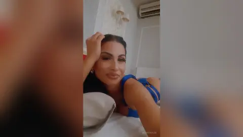 Smiling woman in a blue top relaxing on a white bed in a bright, cozy bedroom setting.