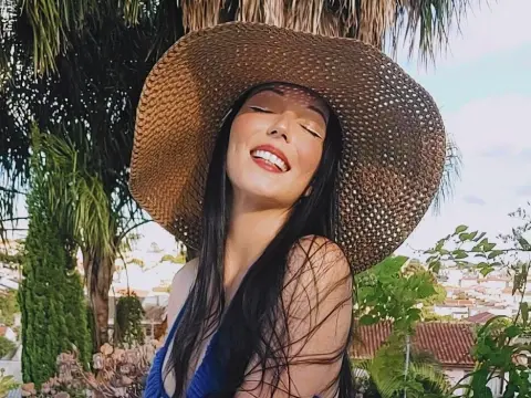 Smiling woman in a wide-brimmed straw hat and blue top enjoying a sunny tropical garden setting.
