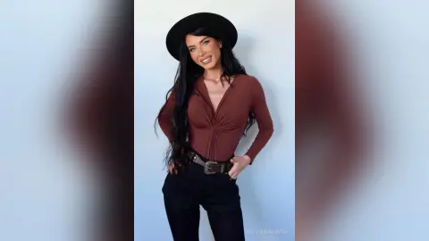 Smiling woman in a brown long-sleeve top, black hat, and belted trousers posing against a light blue background.