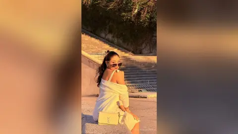 Stylish woman in a white knit outfit and sunglasses sitting on stone stairs during a warm golden hour sunset.