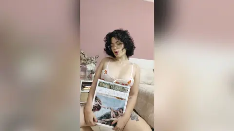 Young woman with curly hair in a white floral bralette holding a magazine in a cozy pink bedroom.