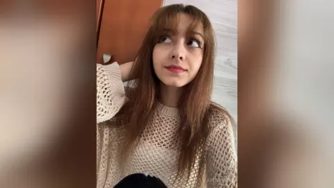 Young woman with long brown hair wearing a cream knit sweater in a bright indoor setting with a dreamy mood.