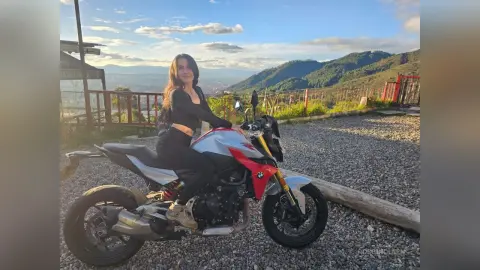 Smiling woman in black casual outfit sitting on a silver sport motorcycle overlooking a scenic mountain valley.