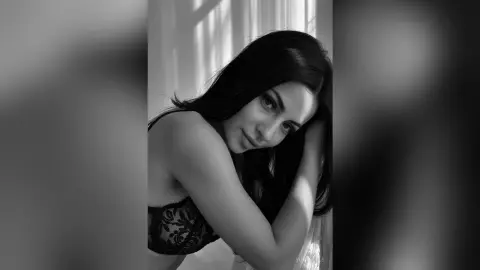 Moody black and white portrait of a woman in black lace lingerie leaning against a sunlit window.
