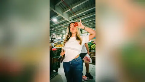 Smiling woman in a white t-shirt and blue jeans holding an apple in a bright grocery store aisle.