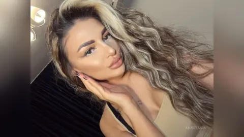 Anastasia poses with her hand against her cheek, showing her long wavy blonde hair and light tan skin.