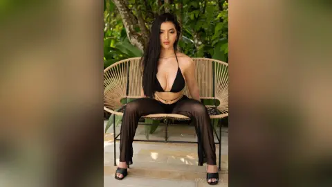 VickyPortman sits on a wicker chair wearing a black bikini top and sheer black trousers against a background of green leaves.