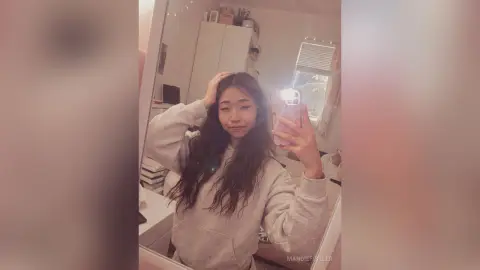 Young woman with long wavy hair wearing a cozy grey sweatshirt taking a casual mirror selfie in a bedroom.