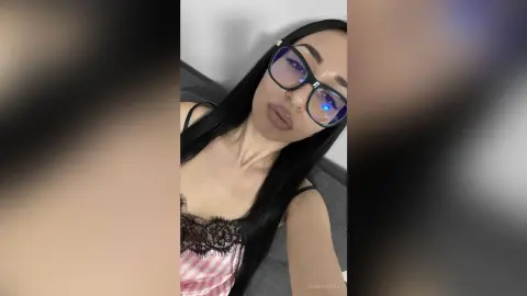 Young woman with long black hair and glasses wearing a pink lace camisole in a relaxed indoor setting.
