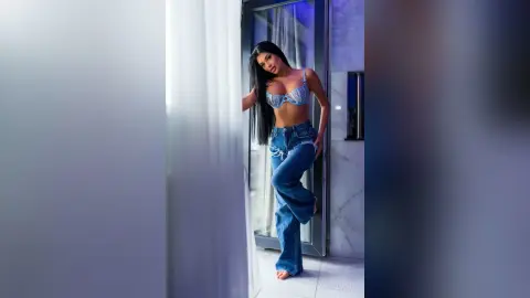 Glamorous woman in a sparkly silver bra and blue denim jeans posing by a bright window in a modern room.