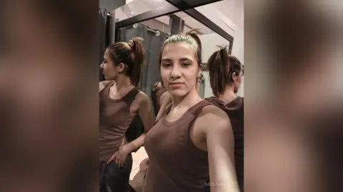 Young woman in a brown tank top taking a mirror selfie in a modern dressing room with a neutral mood.