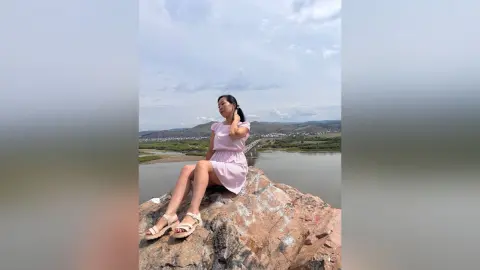 Young woman in a pink summer dress sitting on a rocky cliff overlooking a scenic river valley under a cloudy sky.
