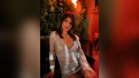 LauraRiveiro — LauraRiveiro poses in a sparkly silver sequin dress with a deep V-neckline while sitting on a dark sofa.
