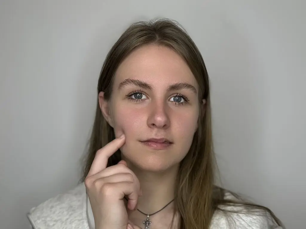 CatherynBuckland looks directly at the camera with long brown hair, wearing a white textured top and a black choker necklace.