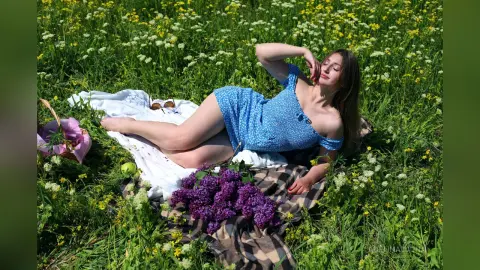 Relaxed woman in a blue floral dress lying on a picnic blanket in a sunny wildflower meadow with purple lilacs.