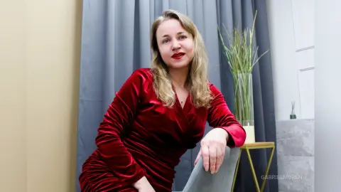 GabrielMoren wears a red velvet wrap dress while sitting in a room with grey curtains and a glass vase of green grass.