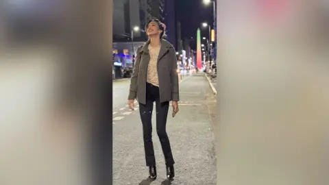 HaleyLyon stands on a city street at night, wearing a grey jacket over a light top and black skinny jeans with black boots.
