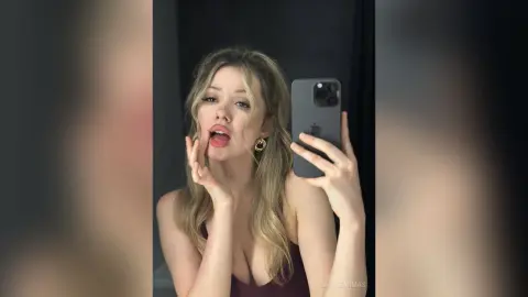 CassieArmas takes a mirror selfie wearing a dark tank top, showing her blonde hair and open mouth while holding her phone.